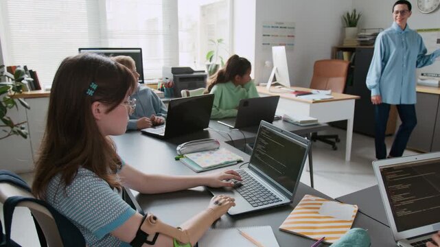 Handheld zoom in shot of young girl with prosthetic arm typing code on laptop during group programming lesson in modern school