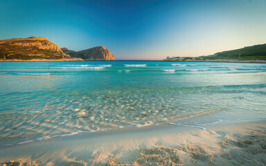 Scenic tropical beach with clear turquoise water gentle waves and rocky cliffs under bright blue sky for travel destination and summer vacation background