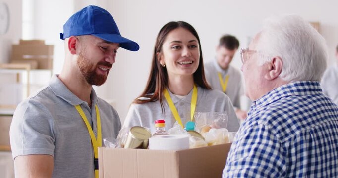 Volunteers in warehouse or community center provide assistance to elderly man by handing him food. Gray-haired man smiles gratefully while taking box of groceries. Concept of helping elderly.