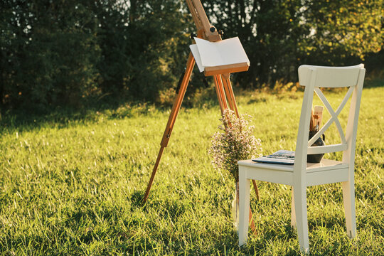 Serene outdoor painting scene. Wooden easel, blank canvas, and white chair on a lush green lawn, bathed in golden sunlight.  Perfect for art, nature, and relaxation themes. - Powered by Adobe