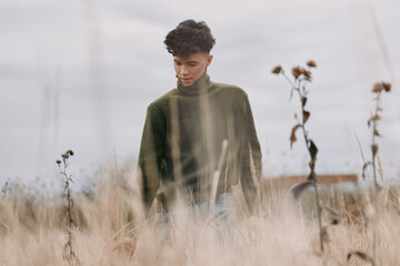 Contemplative young man in a textured green sweater walks through tall dry grass, captured in a...