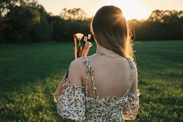 Young woman in floral dress, holding paintbrushes at sunset in a field.  Golden hour light...