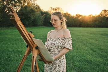 Young woman painting on a meadow at sunset. A beautiful artist in a floral dress standing by her easel, inspired by the golden hour light and the tranquil nature around her. 