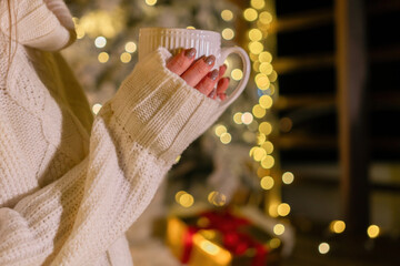 Woman Christmas Coffee - Holding a white mug, wearing a sweater in front of a Christmas tree with lights.