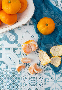 Top View of Tangerines in a White Bowl on Blue and White Tile Background; One Tangerine Out of Bowl on Blue Cloth; One Tangerine Peeled