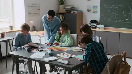Five students collaborating on projects around classroom desk, receiving guidance from female teacher