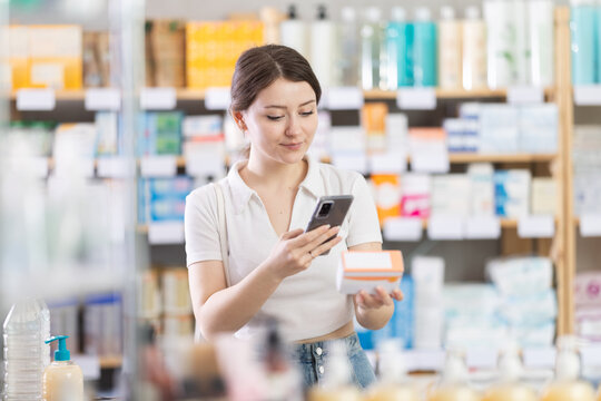 Young woman shopping in pharmacy scanning barcode on box of pills with smartphone, carefully checking dosage, side effects, or contraindications before purchase, ensuring safe self-medication