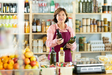 Polite mature female seller kindly offering bottled olive oil in grocery store