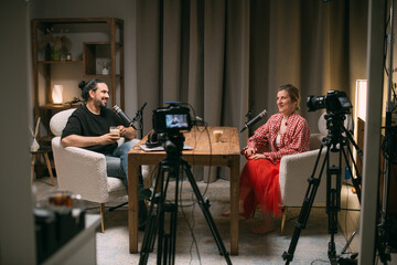Video podcast recording studio. A conversation between a man and a woman sitting in front of microphones and cameras. The host of the podcast is interviewed about the release of a new episode.
