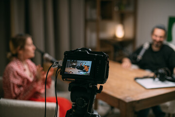 Video podcast recording studio. A conversation between a man and a woman sitting in front of microphones and cameras. The host of the podcast is interviewed about the release of a new episode.