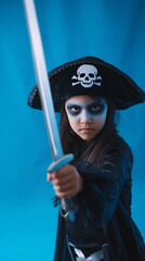 Young hispanic girl in halloween ghost pirate costume with skull hat and toy sword posing playfully against blue studio background. 