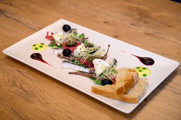 Fine dining seafood starter with sardines, cheese, olives, sprouts, and toast on a white plate with artistic sauces on rustic wooden restaurant table.