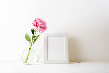 Minimal still life with pink peony in glass vase and blank picture frame on a table against white wall. Mockup