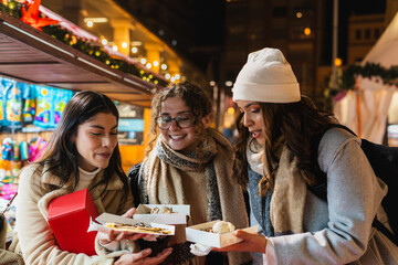 Three friends sharing sweet treats at a festive night christmas market