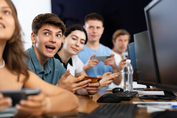 At computer school during class, young guy student sits with mobile phone and listens admiringly to teacher