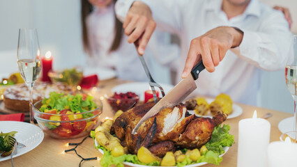 Closeup view of dad cutting roasted turkey for Christmas or Thanksgiving, celebrating traditional holiday with his wife and daughter, having festive dinner at home, copy space