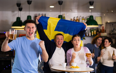 Company of excited young adult sports fans waving flag of Sweden and supporting national team with...