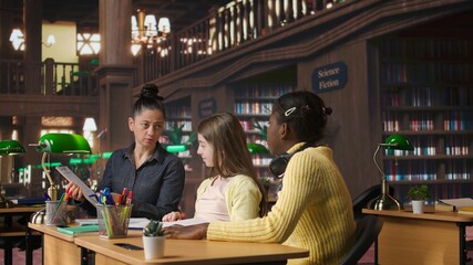Elementary school pupils working with a professor solving exercises for the class homework, developing learning skills at a study desk. Tutor explains the curriculum materials to girls. Camera A.