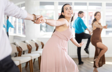 Smiling enthusiastic young dark-haired girl rehearsing upbeat jive dance moves with male partner in sunlit studio. Active hobby concept..