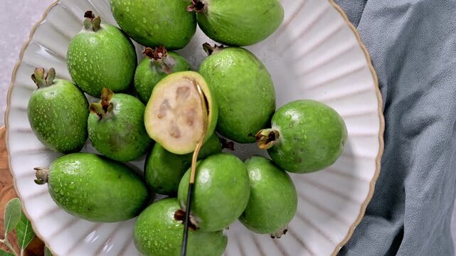 A close-up, top-down view of freshly harvested feijoa fruits, some covered in water droplets, artfully arranged on a fluted white ceramic plate.