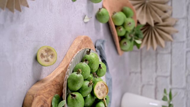 A close-up shot of freshly harvested feijoa fruits, some with glistening water droplets, spilling from a white fluted plate.