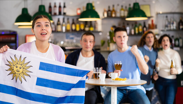 Сompany of emotional young adult sports fans waving flag of Uruguay and supporting favorite team with beer in pub - Powered by Adobe