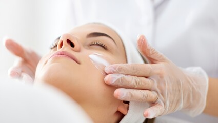 Naklejka premium Woman lying down with eyes closed, receiving a professional facial skincare treatment from a cosmetologist, who is carefully applying a white cream to her cheek