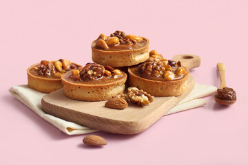 Tasty tarts with nuts and caramel on wooden board against pink background