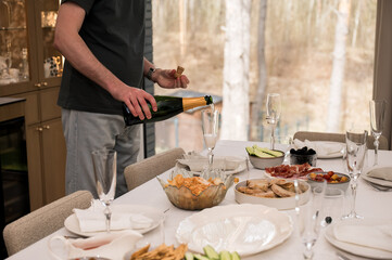 Man pouring champagne into glasses at a festive table set with snacks, bread, and charcuterie during a celebration.