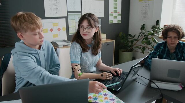Teenage boy helping girl with prosthetic arm with her assignment on laptop during IT class in modern school