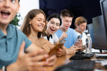 Enthusiastic young girl undergoing training in mobile applications development, sitting at table with computer and using smartphone to test app prototype