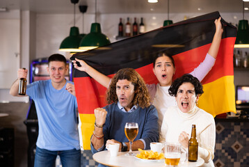 Group of young adult sports fans rooting for favorite team and waving flag of Germany while watching match together in pub