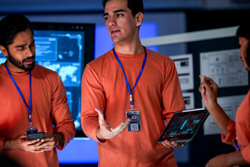 A diverse team of professionals in orange shirts works together in a high tech control room. They are looking at a tablet, discussing a new project or solving a problem.