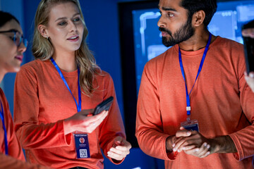 A diverse team of professionals in orange shirts works together in a high tech control room. They are looking at a tablet, discussing a new project or solving a problem.