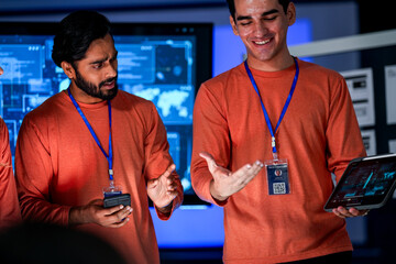 A diverse team of professionals in orange shirts works together in a high tech control room. They are looking at a tablet, discussing a new project or solving a problem.
