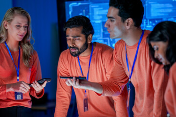 A diverse team of professionals in orange shirts works together in a high-tech control room. They are focused on a smartphone, discussing a new project or solving a problem.