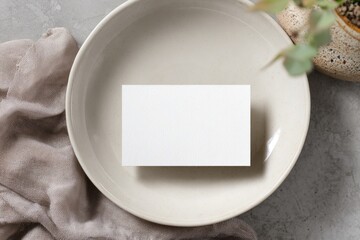 Blank business card on a light beige bowl, nestled amongst a muted taupe fabric draped over a gray concrete surface. A small potted plant sits in the background