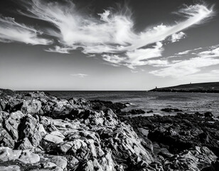 Dramatic black and white landscape of a rugged rocky coastline under a sky filled with wispy clouds, with distant ocean and land formations.