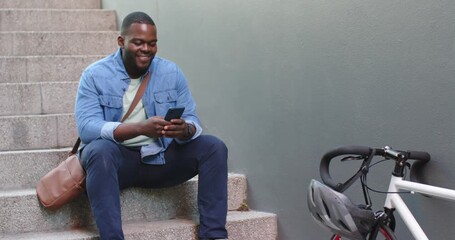 African American man sitting on steps by grey wall tapping smartphone reading messages with bike - Powered by Adobe