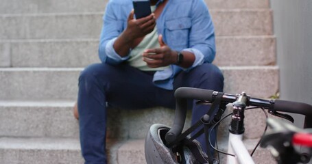 African American man tapping smartphone after alert making call gesturing on stairs by bike helmet - Powered by Adobe