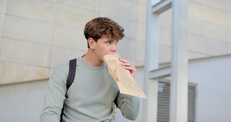 Man wearing backpack outside building pulling paper bag open, nibbling sandwich and checking plaza - Powered by Adobe