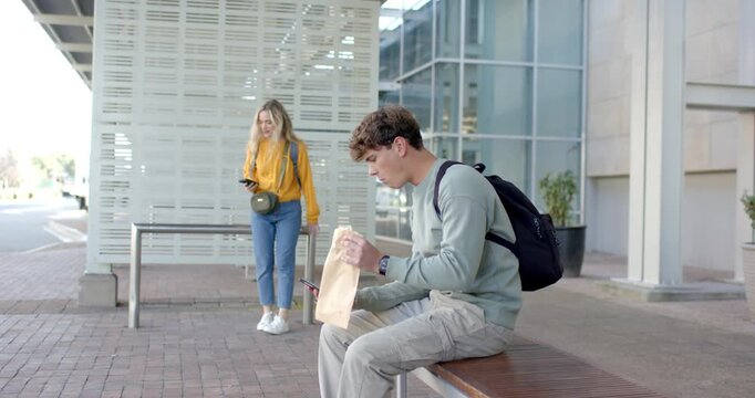 Man holding phone at shelter bench as woman waiting triggers opening sandwich and sipping drink