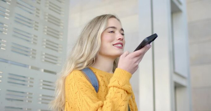 Female wearing sweater and backpack raising phone to mouth and speaking into assistant at building