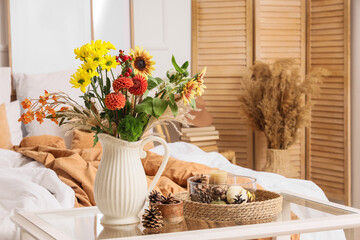 Vase with autumn flowers and fir cones on table in light bedroom