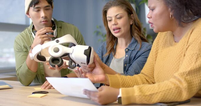Diverse coworkers gathering at office table, examining sheet, passing headset, evaluating VR design