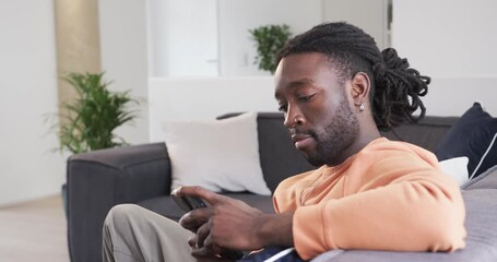 After reading message, African American man scrolling smartphone and speaking reply on grey couch - Powered by Adobe