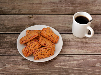 Plate with a pile of fresh homemade shortbread cookies with fruit jam on a wooden table, next to it a cup of hot black coffee.