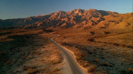Aerial view of a winding desert road leading towards a majestic mountain range bathed in the warm hues of sunset.  The landscape is arid, with sparse vegetation and textured earth tones