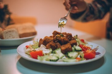 A plate topped with meat and fresh vegetables, a hand using a fork to drizzle dressing. Breads are visible in background, creating soft lighting