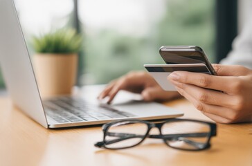 A person is using a smartphone and credit card in front of an open laptop on a wooden table with eyeglasses and a potted plant in the background